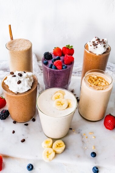 Six protein shakes arranged on a marble board, topped with berries, banana slices, peanut butter, chopped peanuts and whipped cream.