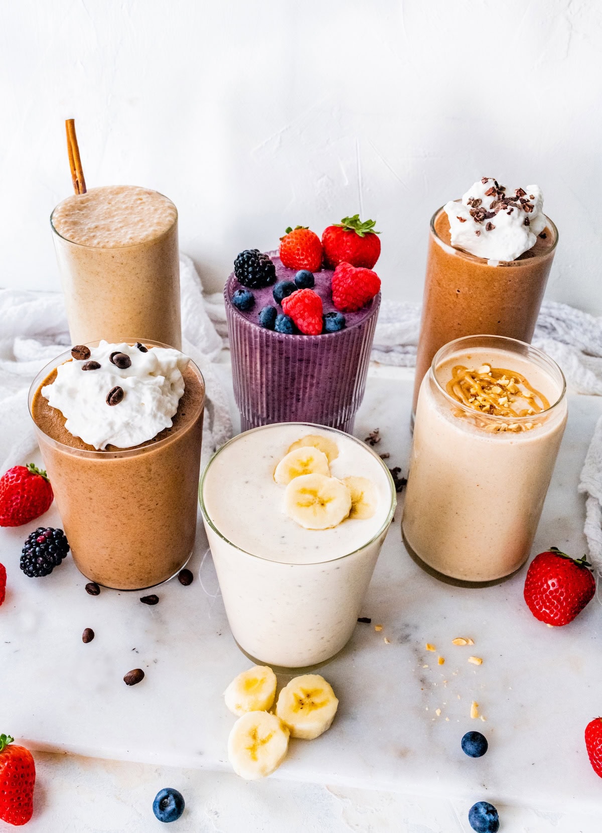 Six protein shakes arranged on a marble board, topped with berries, banana slices, peanut butter, chopped peanuts and whipped cream.