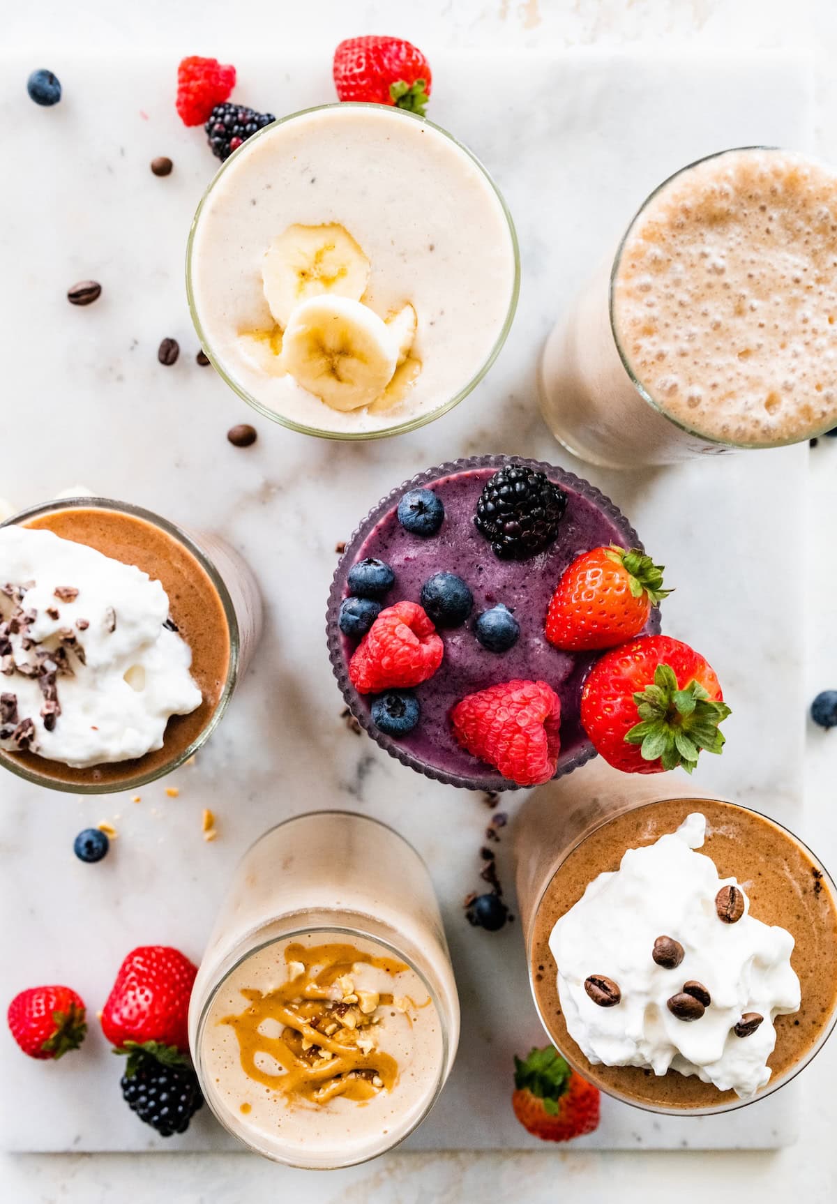 Six protein shakes in glasses with assorted toppings, including fresh berries, banana slices and whipped cream.