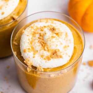 Overhead close-up of pumpkin mousse topped with whipped topping and graham cracker crumbs beside a small orange pumpkin.