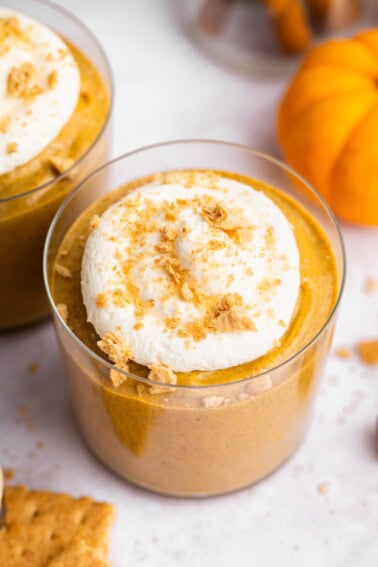 Overhead close-up of pumpkin mousse topped with whipped topping and graham cracker crumbs beside a small orange pumpkin.
