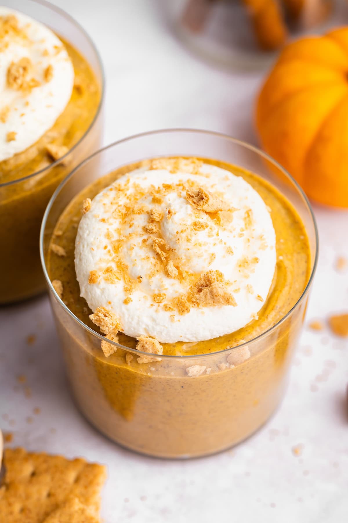 Overhead close-up of pumpkin mousse topped with whipped topping and graham cracker crumbs beside a small orange pumpkin.