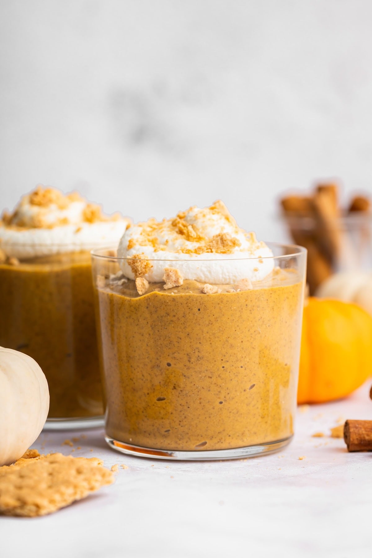 Two glasses of pumpkin mousse topped with whipped topping and graham cracker crumbs, with mini pumpkins nearby.
