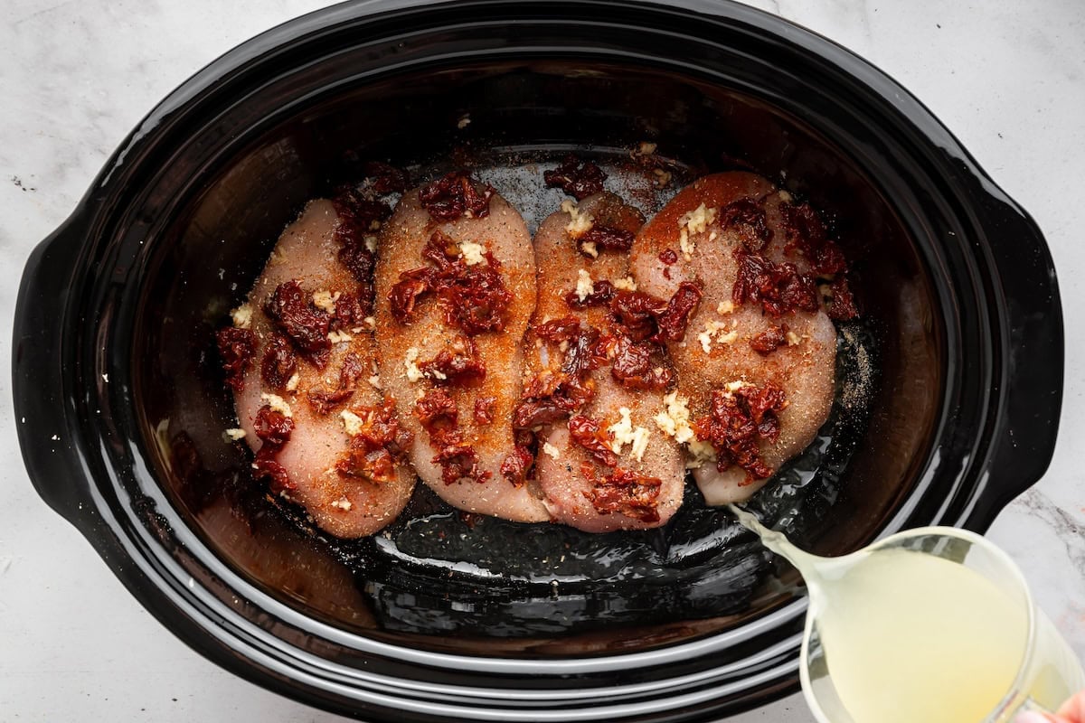 Seasoned chicken breasts topped with minced garlic and sun-dried tomatoes in the slow cooker with broth being poured over everything.