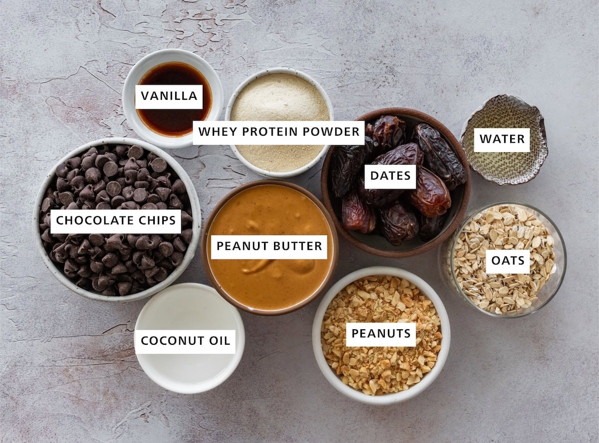 Ingredients measured out for snickers protein balls include: peanut butter, peanuts, oats, water, dates, whey protein powder, vanilla, chocolate chips, and coconut oil.