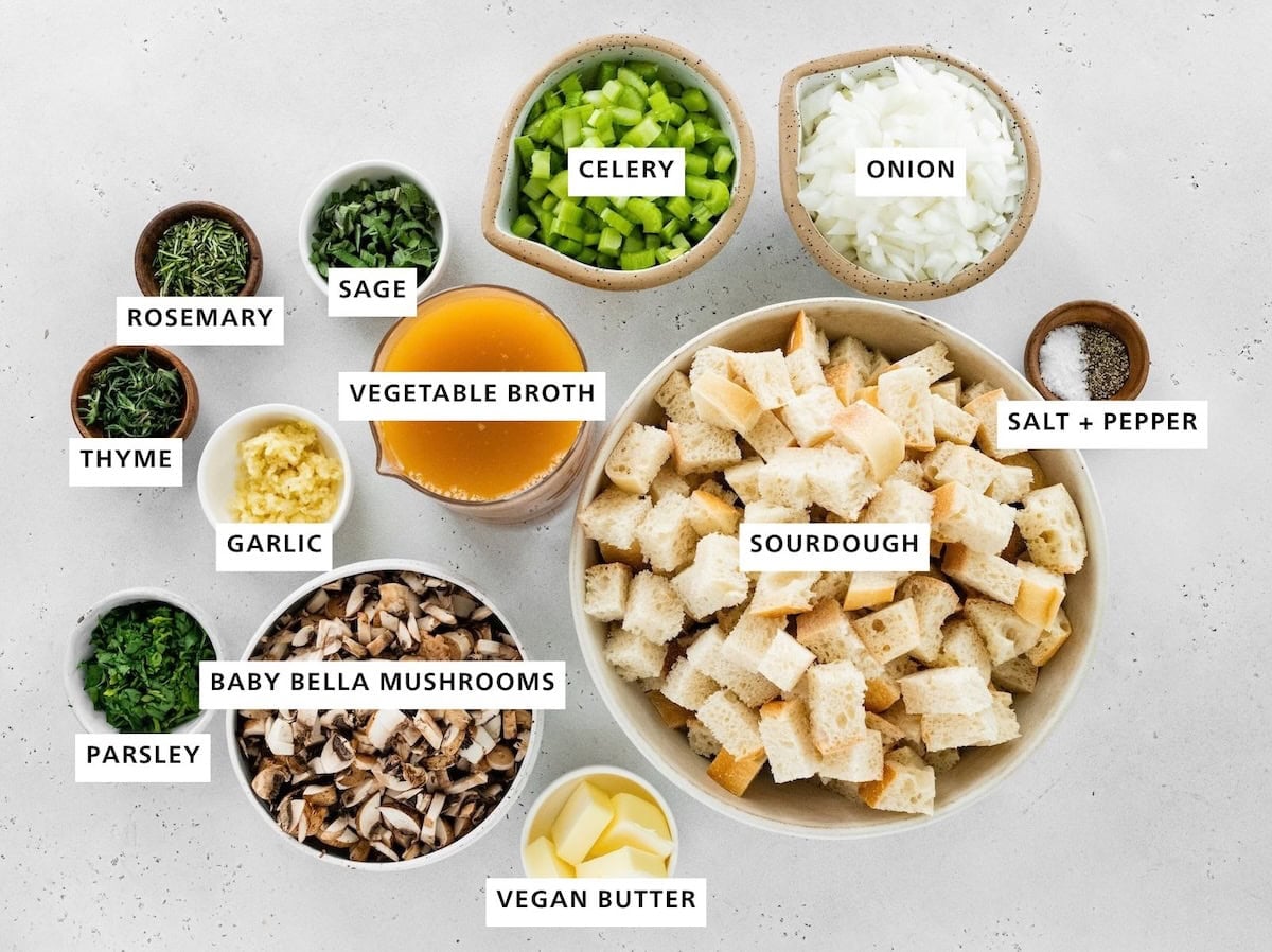 Ingredients measured out to make Sourdough Stuffing: salt, pepper, onion, sourdough, celery, vegetable broth, vegan butter, sage, baby bella mushrooms, garlic, rosemary, thyme and parsley.