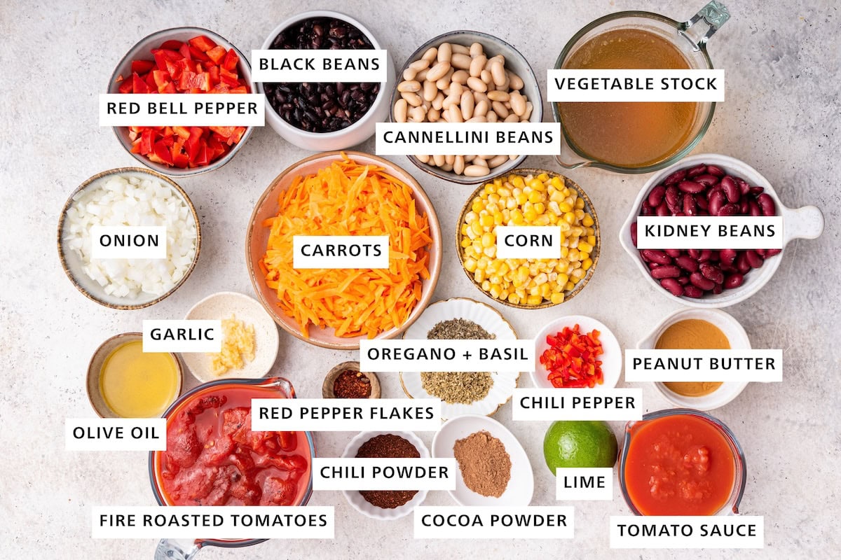Ingredients measured out to make Easy Vegetarian Chili: fire roasted tomatoes, olive oil, onion, red bell pepper, garlic, red pepper flakes, carrots, black beans, chili powder, oregano, cocoa powder, basil, corn, cannellini beans, lime, chili pepper, tomato sauce, peanut butter, kidney beans and vegetable stock.